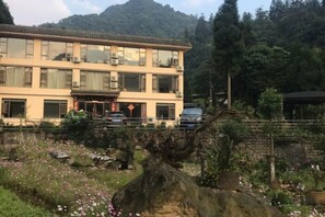 A multi-story building with a traditional architectural style, surrounded by a landscaped garden with flowering plants and a large rock feature. - Mount Emei Feng Lin Yue Hotel (Emeishan City)