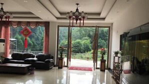 A spacious living room with a large flat-screen TV, a sofa, and a coffee table. There is a large window with curtains and a view of greenery outside.