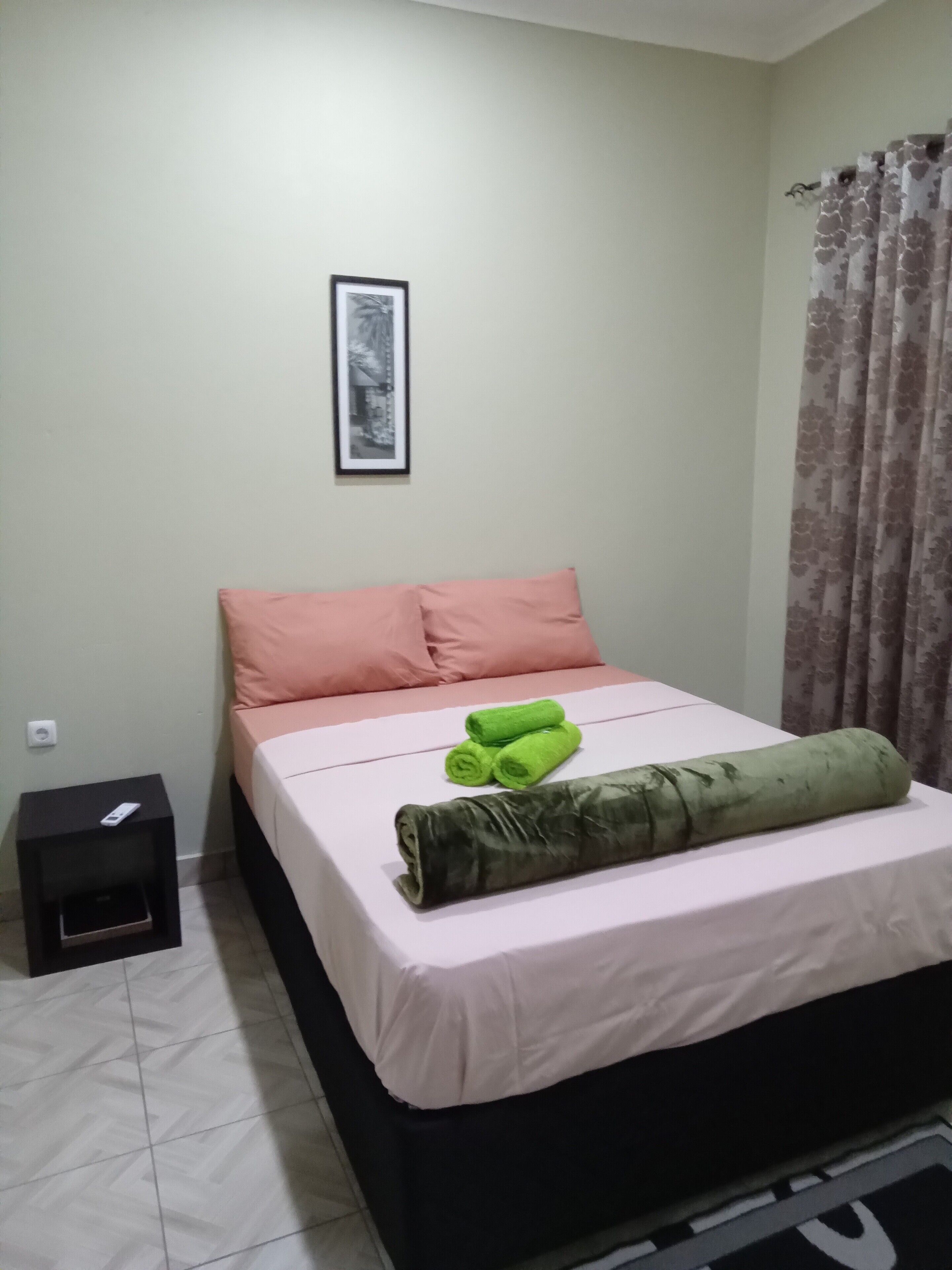 Une chambre à coucher avec un lit, des oreillers, une table de chevet, un tableau encadré au mur et un rideau.