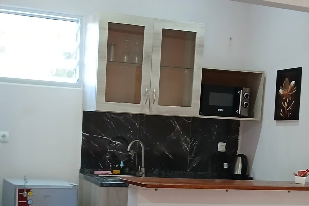 A compact kitchen with a microwave, a sink, and cabinets. There is a chair with a cushion in the foreground.