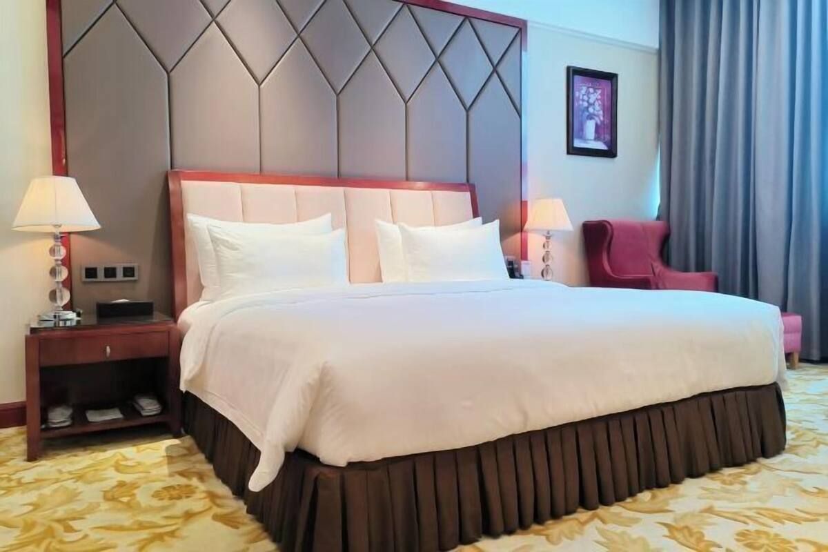 A hotel room with a large bed, a wooden headboard, two bedside lamps, a red chair, and a framed picture on the wall.