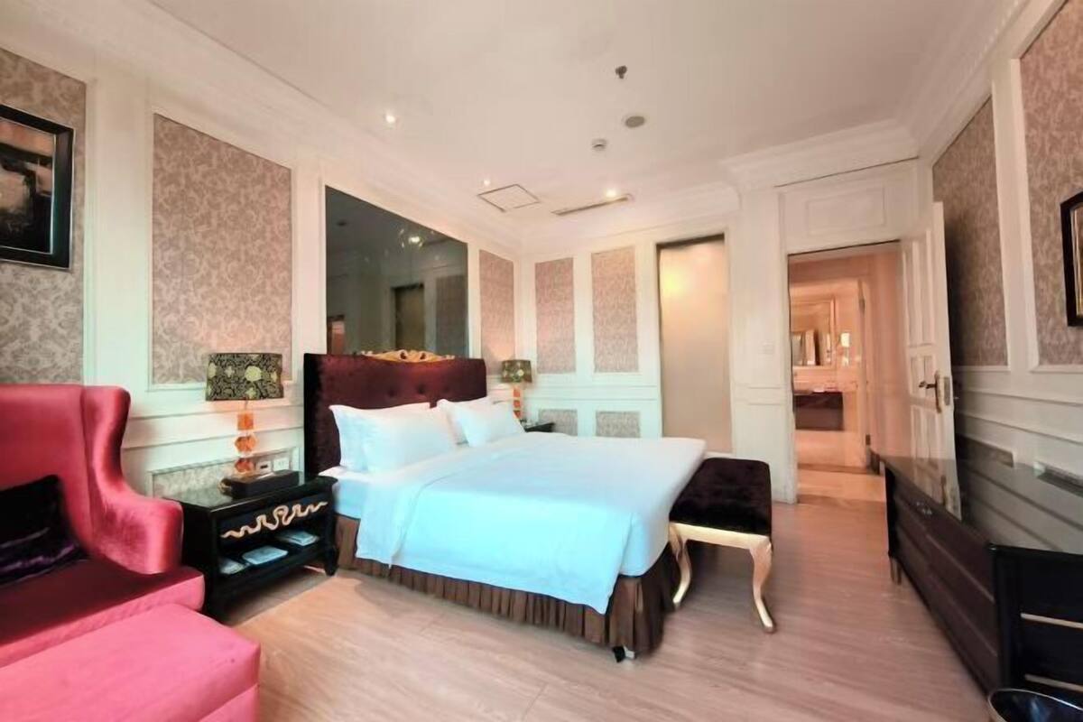 A bedroom with a large bed, a red armchair, a black nightstand, and a mirror on the wall.