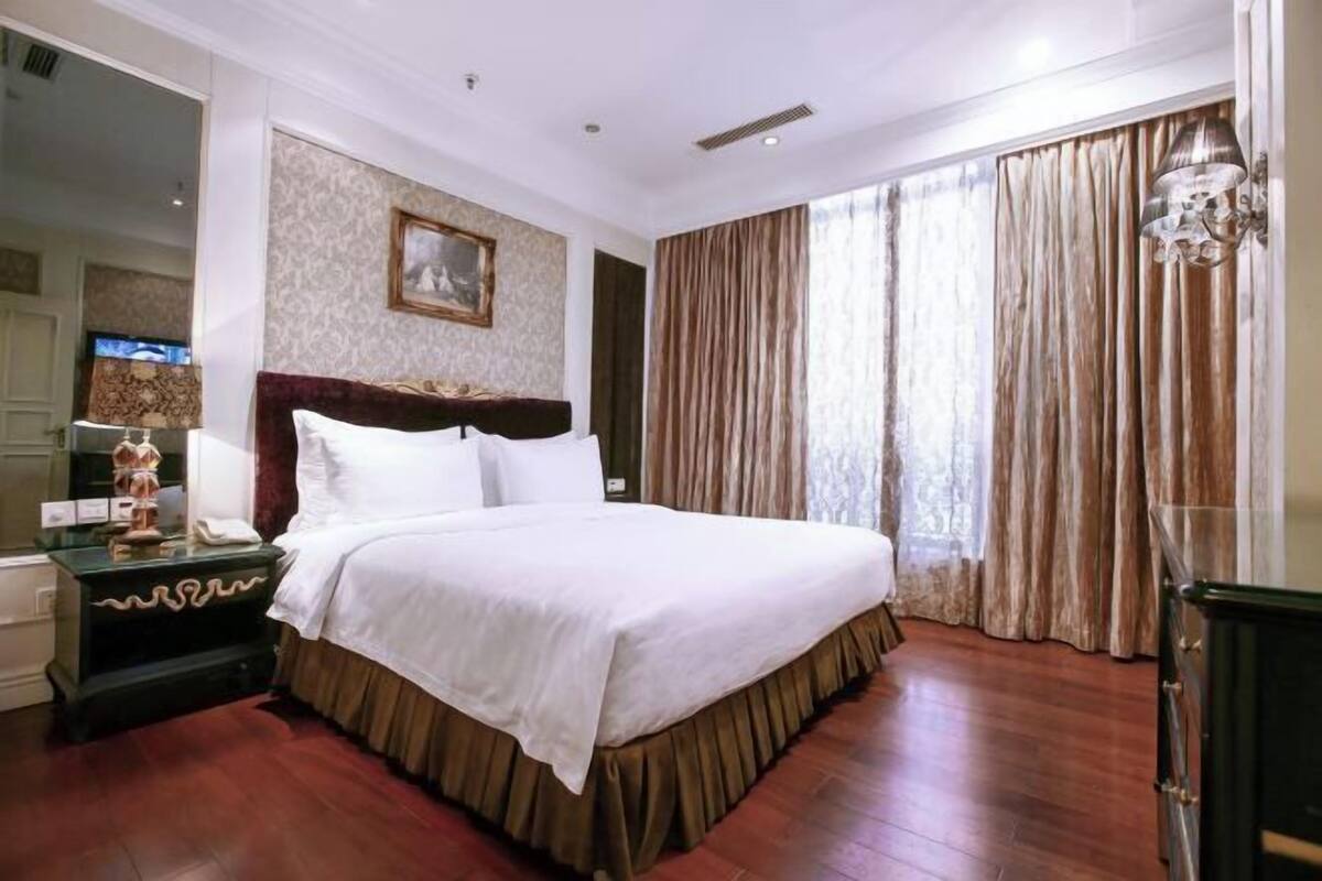 A hotel room with a large bed, a nightstand with a lamp, a mirror, and a view of the outdoors through curtains.