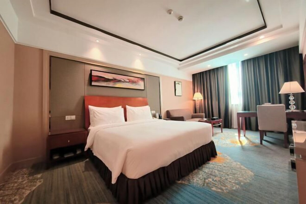 A hotel room with a large bed, a desk, a chair, a lamp, and a painting on the wall.