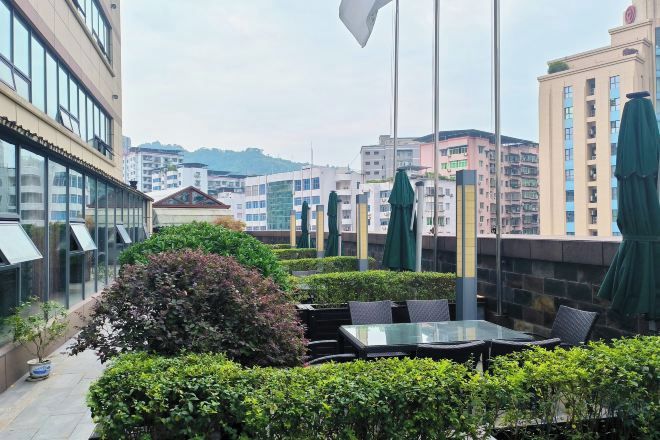 An outdoor seating area with tables and chairs, surrounded by greenery and overlooking a cityscape with buildings.