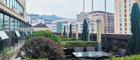 An outdoor seating area with tables and chairs, surrounded by greenery and overlooking a cityscape with buildings.
