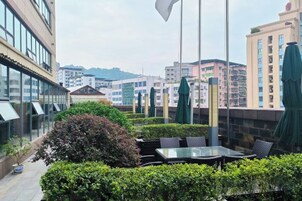 An outdoor seating area with tables and chairs, surrounded by greenery and overlooking a cityscape with buildings.