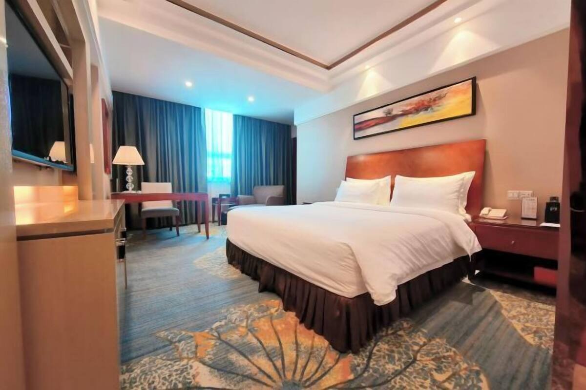 A hotel room with a large bed, a desk with a lamp, a chair, and a window with curtains.