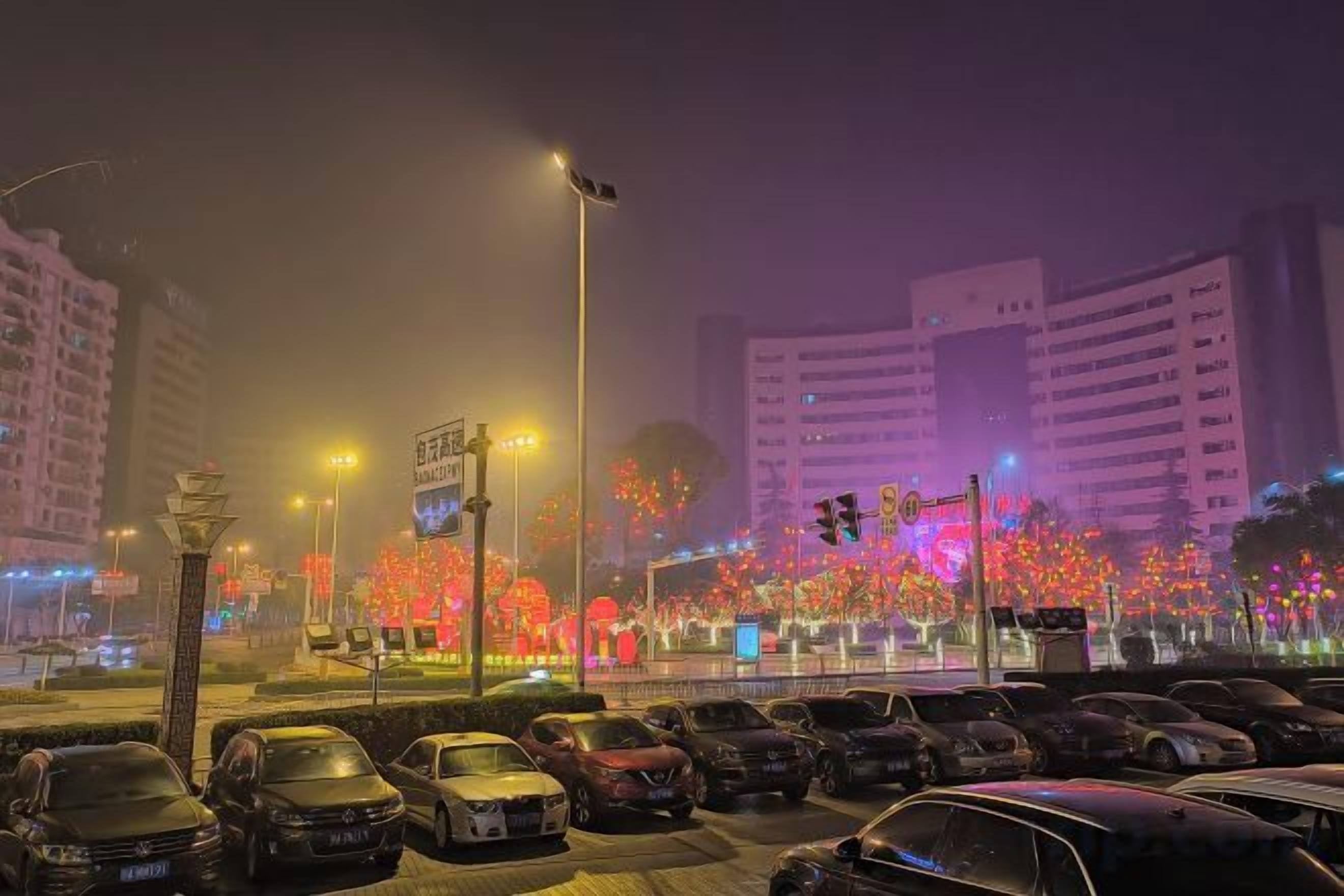 A nighttime cityscape with illuminated buildings and a parking area.
