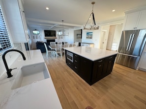 A modern kitchen with a central island, stainless steel appliances, and a dining area with white furniture. - OCEAN BLOCK - BRAND NEW HOME W HEATED POOL AND ELEVATOR (Surf City)