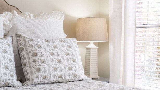Un lit avec des coussins à motifs, une lampe de chevet et une fenêtre avec des stores.