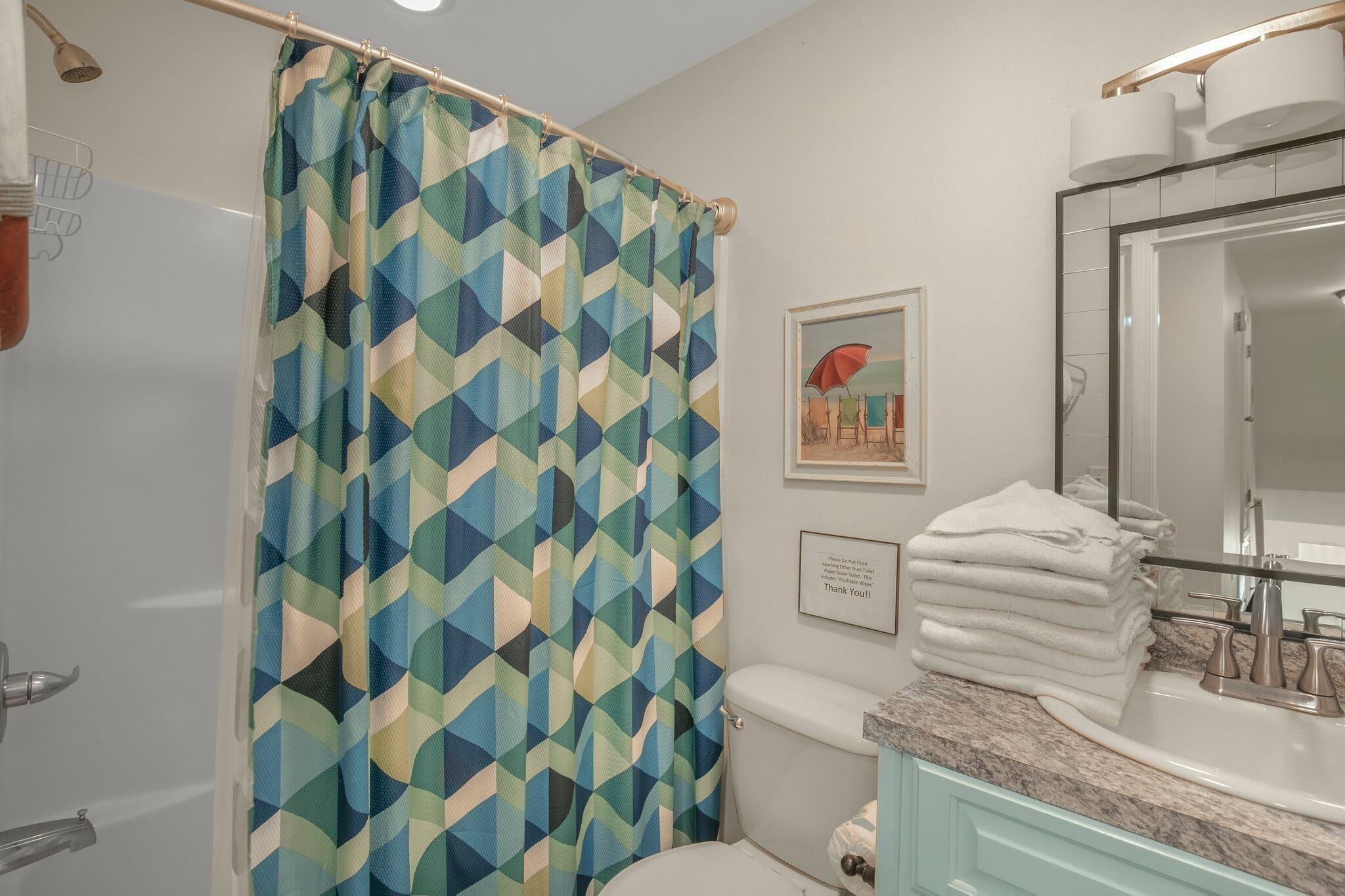 A bathroom with a geometric patterned shower curtain, a toilet, a sink with a marble countertop, and a framed picture on the wall.