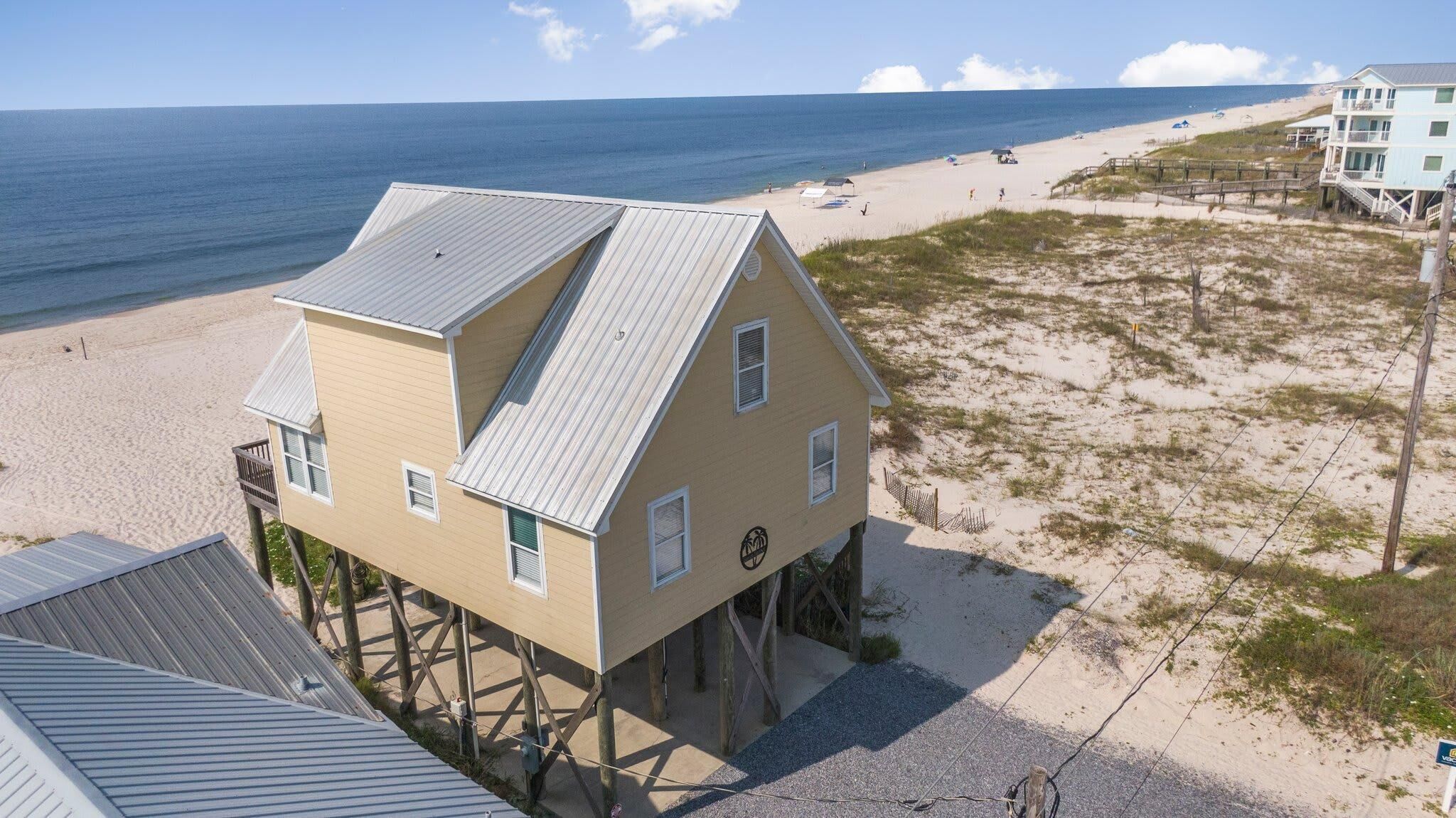 Uma casa de praia de dois andares com telhado de metal, elevada em pilares, localizada numa praia arenosa com vista para o oceano.