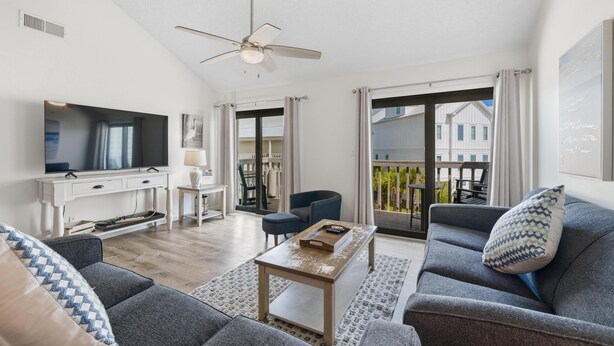 A modern living room with a flat-screen TV, a ceiling fan, a dining area with a table and chairs, and a balcony with a view of houses. - Cotton Bayou 7H (Orange Beach)