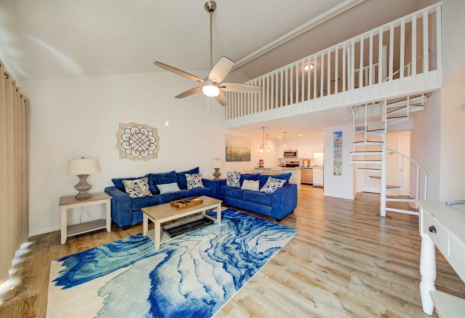 Un salon spacieux avec un canapé bleu, une table basse en bois et un grand tapis bleu. Un ventilateur de plafond et un escalier sont visibles à l’arrière-plan.
