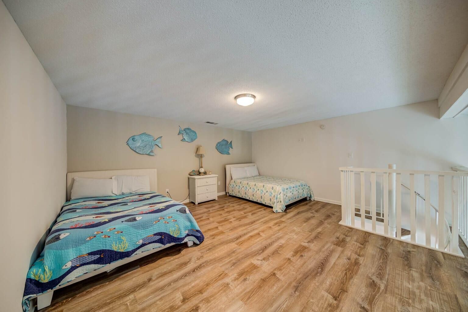 Un dormitorio con suelo de madera, dos camas, una mesita de noche, un aparador y una barandilla blanca.