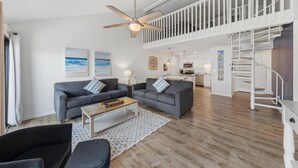 A spacious living area with a high ceiling, a ceiling fan, a staircase, and a kitchen in the background. - Cotton Bayou 7H (Orange Beach)