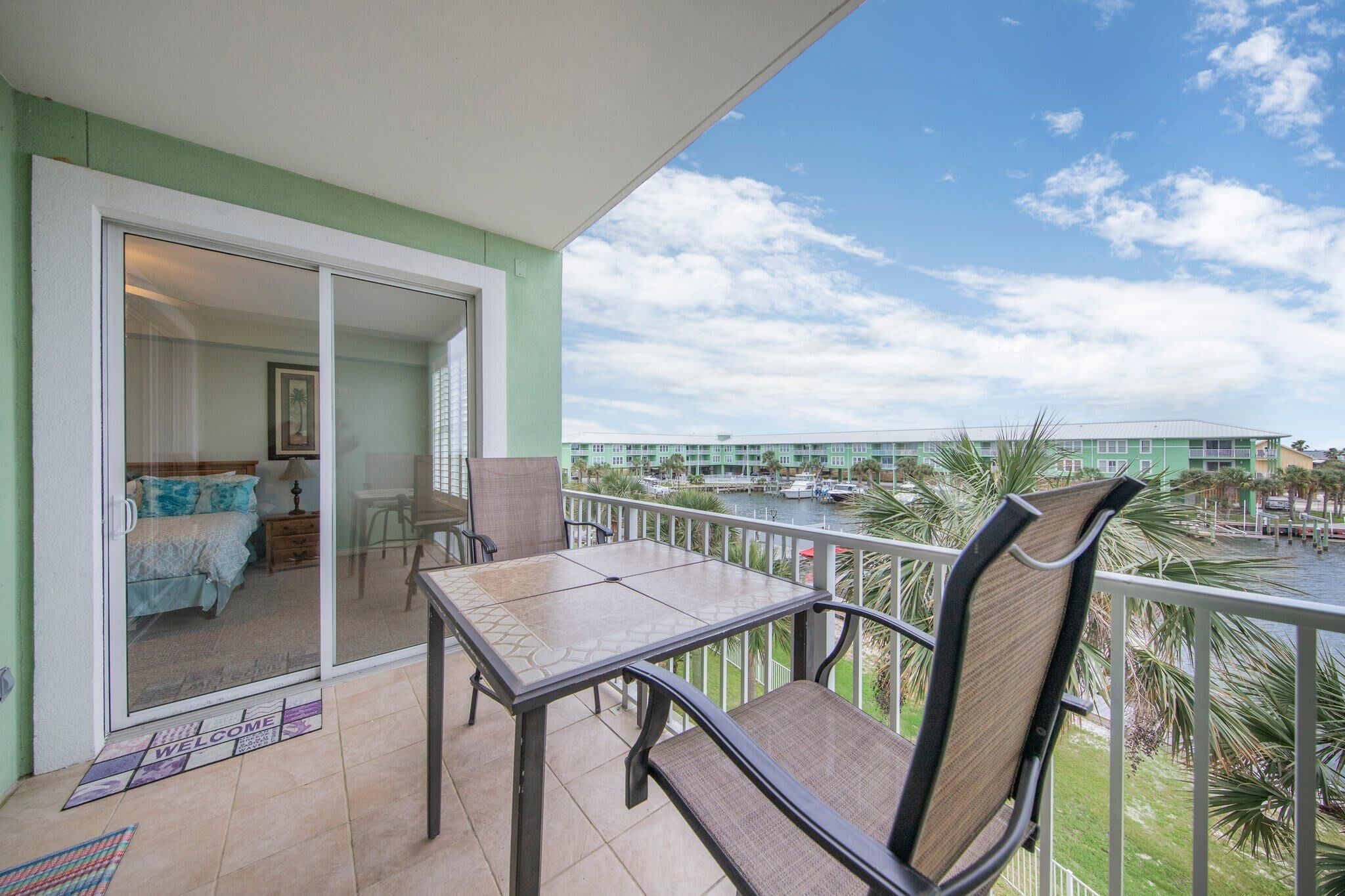 A balcony with a table and chairs, a sliding glass door leading to a bedroom, and a view of a marina with boats.