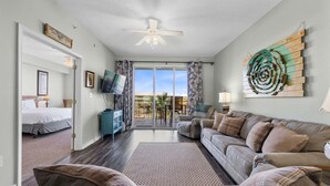 A living room with a sofa, a television, a ceiling fan, and a view of the outdoors.