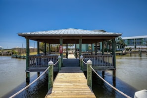Une jetée en bois mène à un belvédère couvert doté de balustrades, qui surplombe l’eau, avec des bâtiments visibles en arrière-plan.