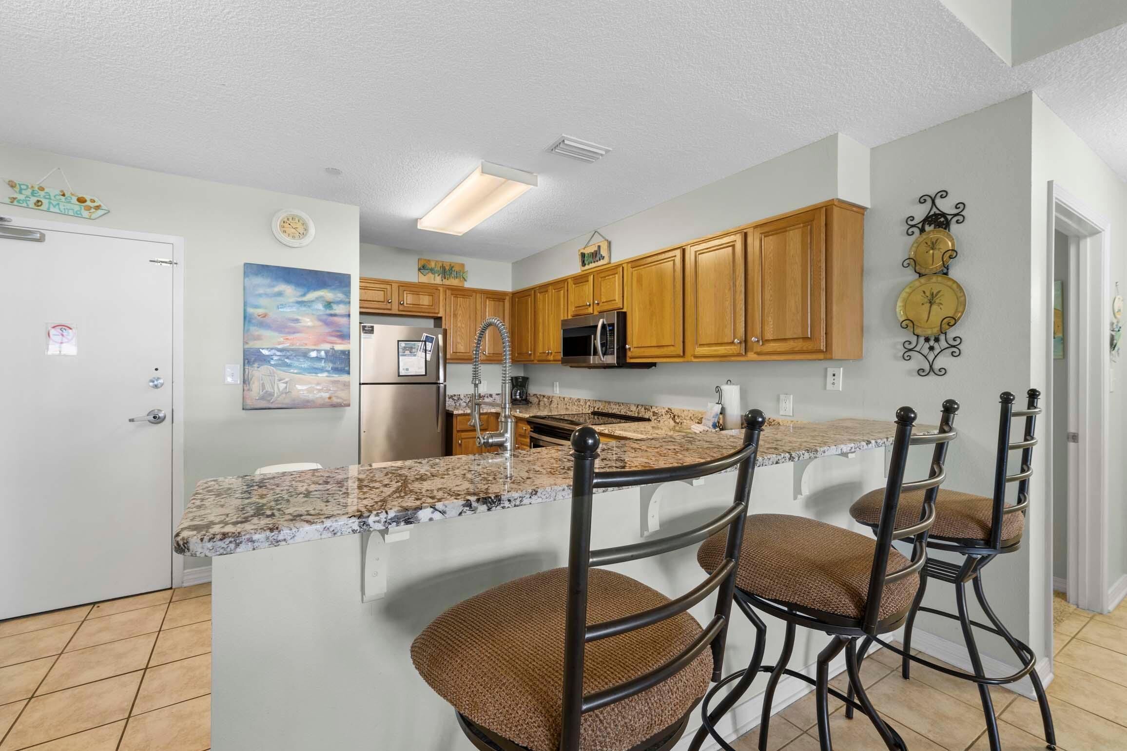 A kitchen with wooden cabinets, granite countertops, a stainless steel refrigerator, and a bar area with stools.
