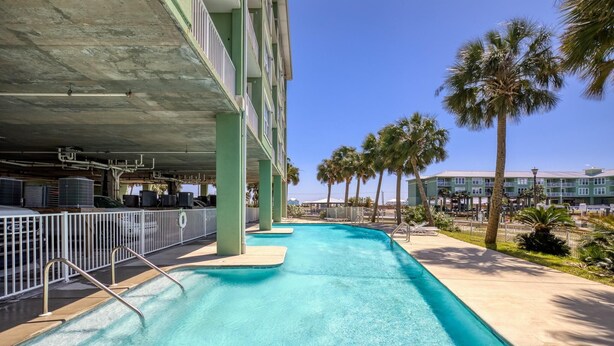 A swimming pool with metal railings, surrounded by palm trees and a building with balconies.