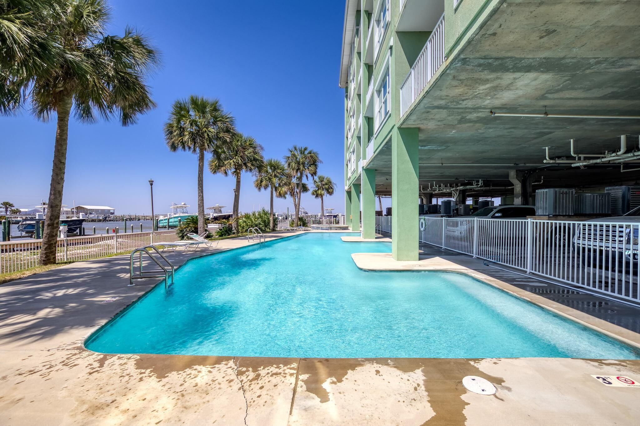 A swimming pool with a concrete deck, surrounded by palm trees and a parking garage.