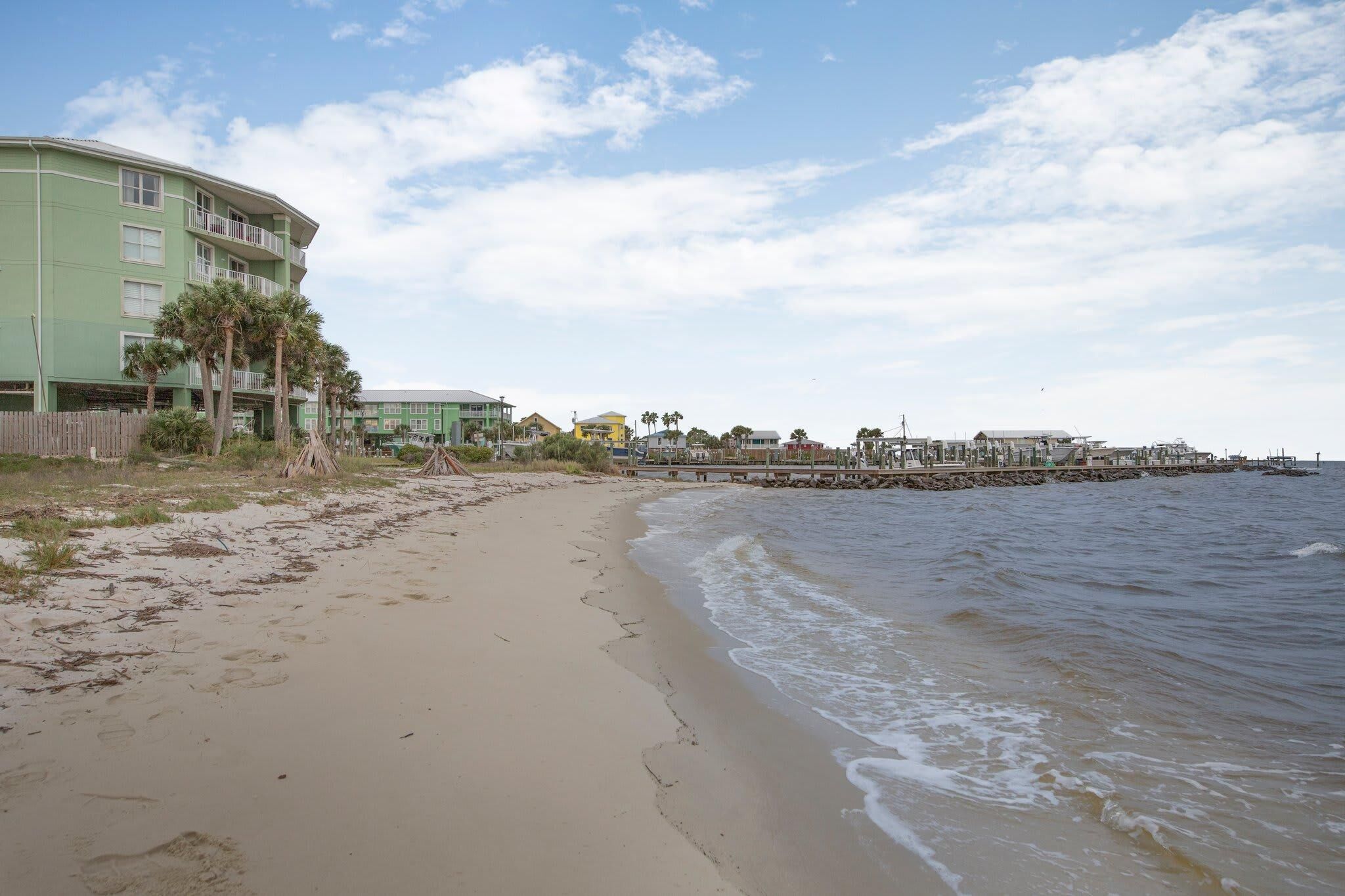 A beach with a wooden pier extending into the water, a green building with balconies, and a clear sky.