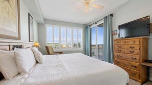A hotel room with a large bed, a ceiling fan, a TV, a wooden dresser, and a balcony with a view.