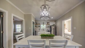 A dining area with a white table and chairs, a chandelier, and a kitchen in the background. - The Rookery 2202 (Gulf Shores)