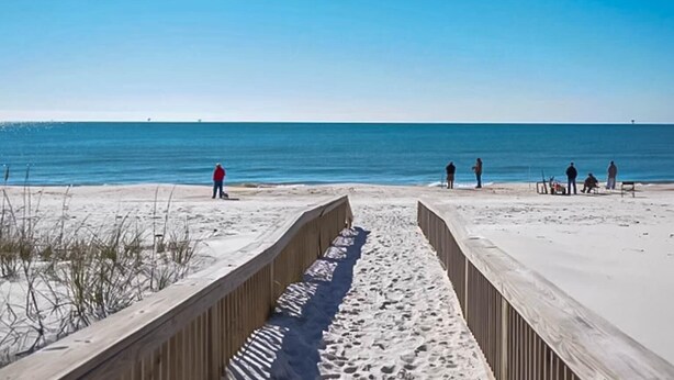 A beach with a wooden walkway leading to the water, people walking, and a clear blue sky. - The Rookery 2202 (Gulf Shores)