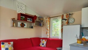 A cozy living room with a red sofa, a small table, and a window with curtains. - Mobile home rental (Agde)