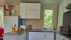 A compact kitchen with white cabinets, a sink, and a window providing a view of greenery outside. - Mobile home rental (Agde)