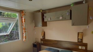 A bedroom with a bed, a window with curtains, a bedside table, and a wall-mounted shelf with decorative items. - Mobile home rental (Agde)