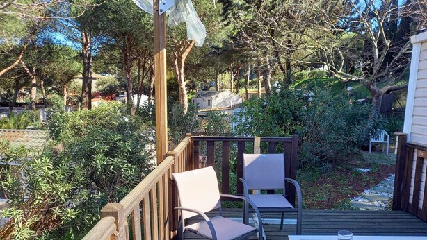 A wooden deck with two chairs and a table, surrounded by lush greenery and trees. - Mobile home rental (Agde)