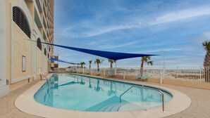 A swimming pool with a curved edge, surrounded by a brick walkway. There are lounge chairs and a shaded area with a blue canopy.
