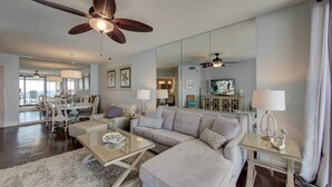 A spacious living room with a large sectional sofa, a glass coffee table, and a dining area with a white table and chairs. There is a ceiling fan and a large mirror reflecting the room. - Pelican Pointe 1505 (Orange Beach)