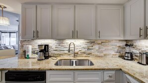 A modern kitchen with white cabinets, a granite countertop, a built-in dishwasher, and a stone backsplash. - Pelican Pointe 1505 (Orange Beach)