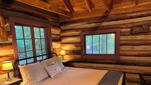 Ein Schlafzimmer in einer Blockhütte mit einem Bett, zwei Fenstern, einer Lampe und einem hölzernen Kopfstück.
