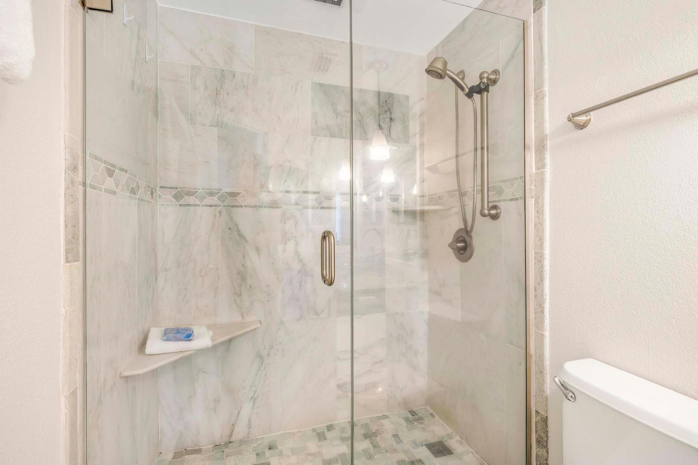 A glass-enclosed shower with a marble floor and a white bathtub visible to the right.