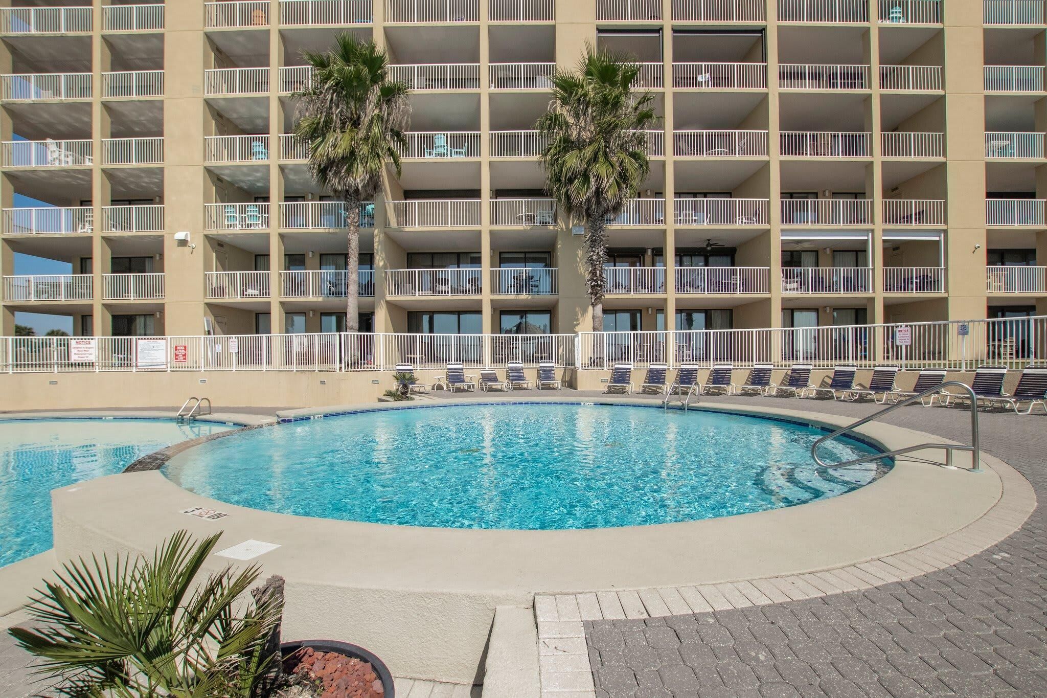 A swimming pool with lounge chairs and palm trees, surrounded by a multi-story building with balconies.