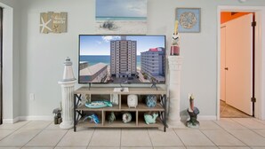 A living room with a television displaying a beach scene, a shelf with decorative items, and a door leading to another room. - Gulf Tower 3C (Gulf Shores)
