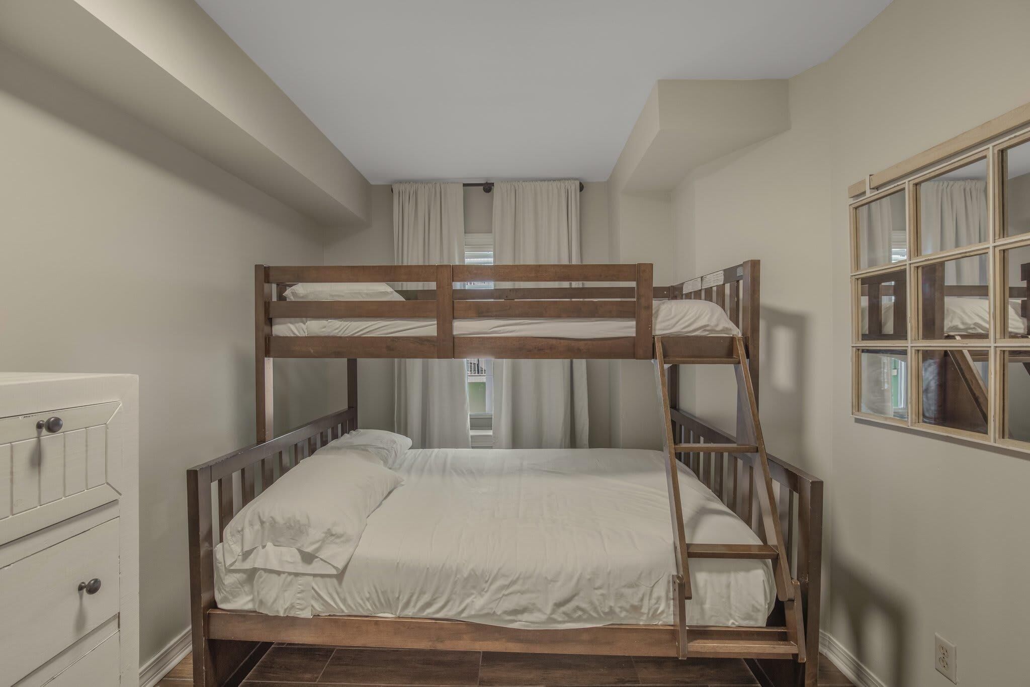 Una cama individual superpuesta en una habitación con un aparador, una ventana con cortinas y un espejo en la pared.