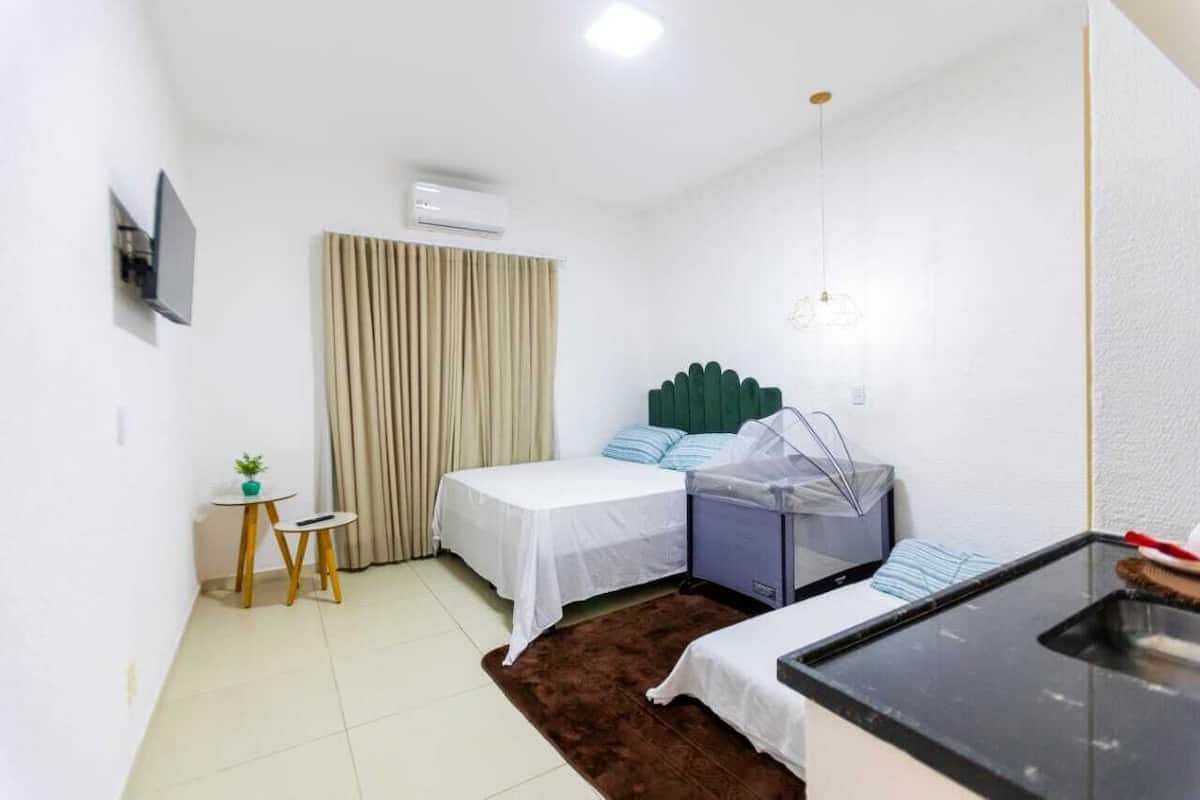 Habitación de hotel pequeña con dos camas, una mesita con una planta, una kitchenette y una ventana con cortinas.