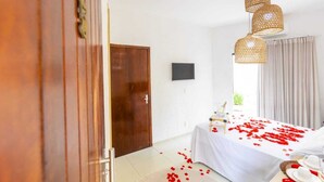 Habitación de hotel con una cama cubierta de pétalos de rosa, un armario de madera, un televisor de pantalla plana y una lámpara en el techo.