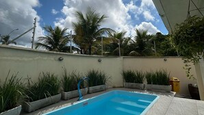 Una piscina rectangular con superficie de agua azul cristalina, rodeada de plantas en macetas y un suelo de baldosas.