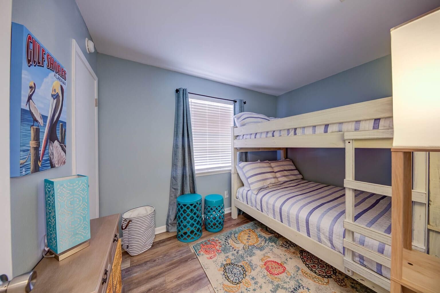 A bedroom with a bunk bed, a window with curtains, a framed picture on the wall, and a patterned rug.