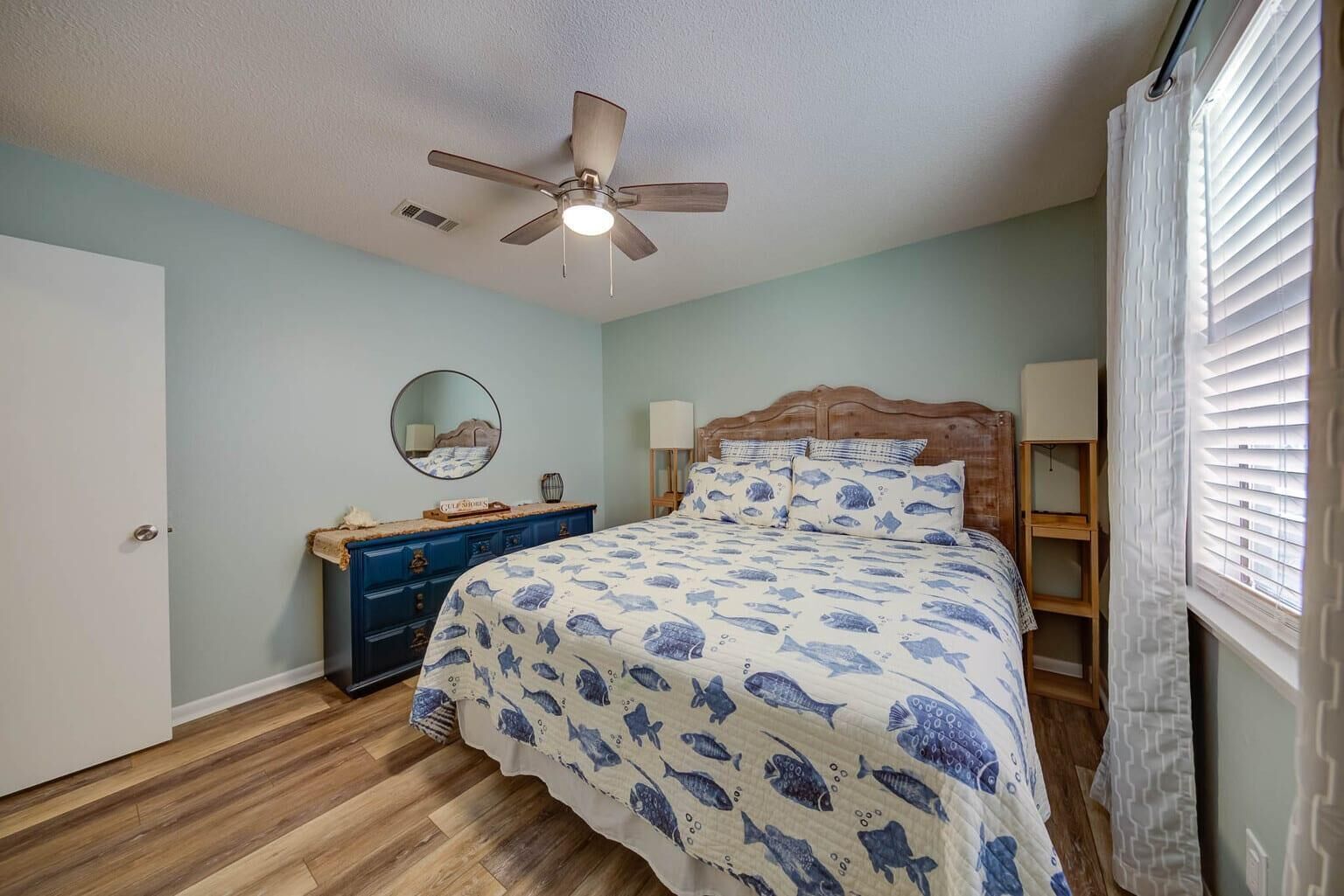 Une chambre à coucher comprenant un lit, une commode, un ventilateur de plafond et une fenêtre avec des stores.