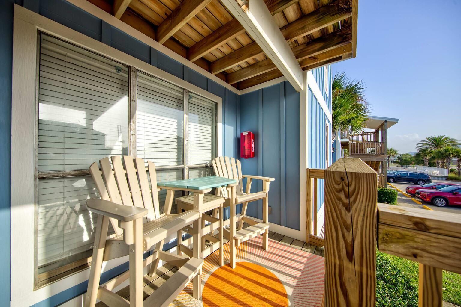 A covered outdoor seating area with wooden chairs and a table, a wooden deck, and a view of parked cars and palm trees.