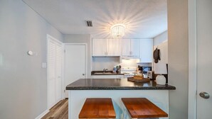 Cocina compacta con armarios blancos, encimera de granito y dos taburetes de madera.